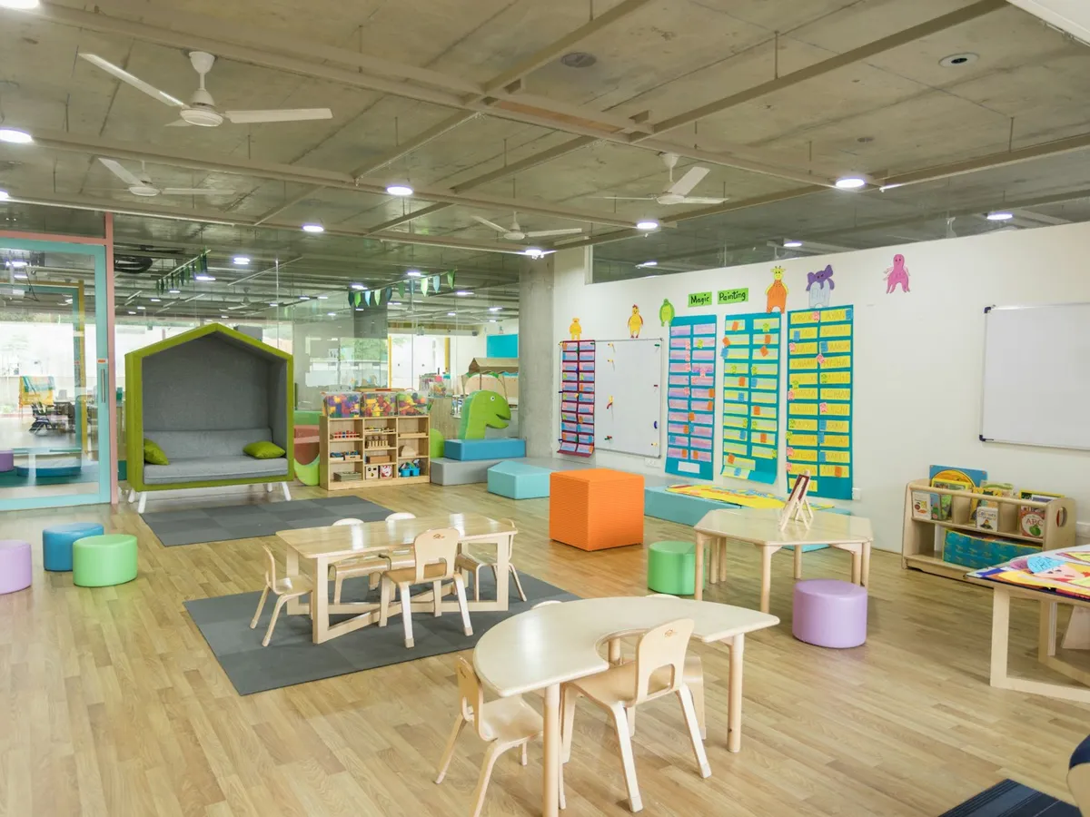 Bright childcare room with small tables, chairs, and play areas without children present